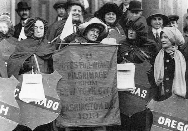 General Rosalie Jones holding up the hike's official banner (made before road conditions forced a start in Newark, rather than New York City, where the kickoff rally was held), with Colonel Ida Craft to the left holding up a shield with the name of the equal-suffrage State of Colorado. Elizabeth Freeman is on the far right, holding up the name of Oregon, another State where women have the same voting rights as men.